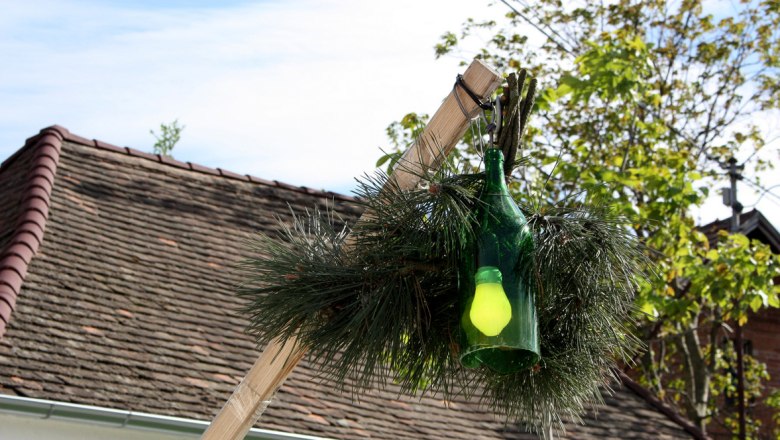 Eine grüne Flasche mit einer gelben Glühbirne hängt an einem Holzstab, umgeben von Tannenzweigen, vor einem Dach und Bäumen.