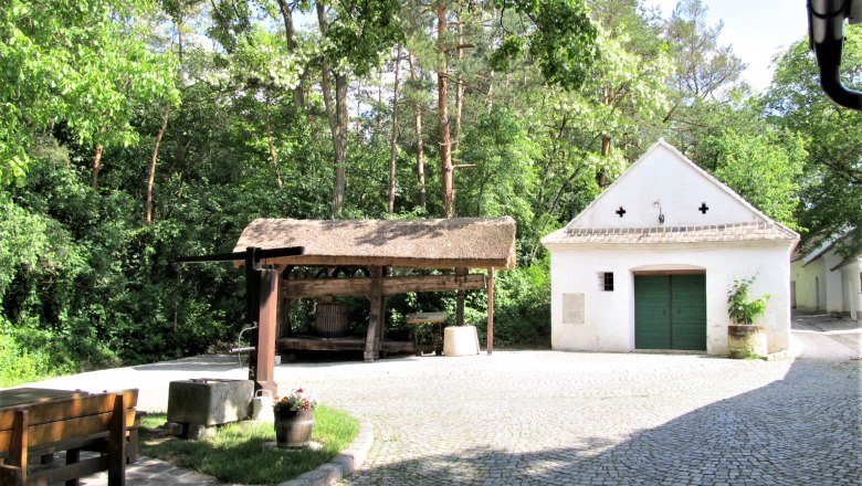 Ein traditioneller Weinkeller mit einer alten Weinpresse und einem kleinen wei&szlig;en Geb&auml;ude im Hintergrund, umgeben von B&auml;umen.