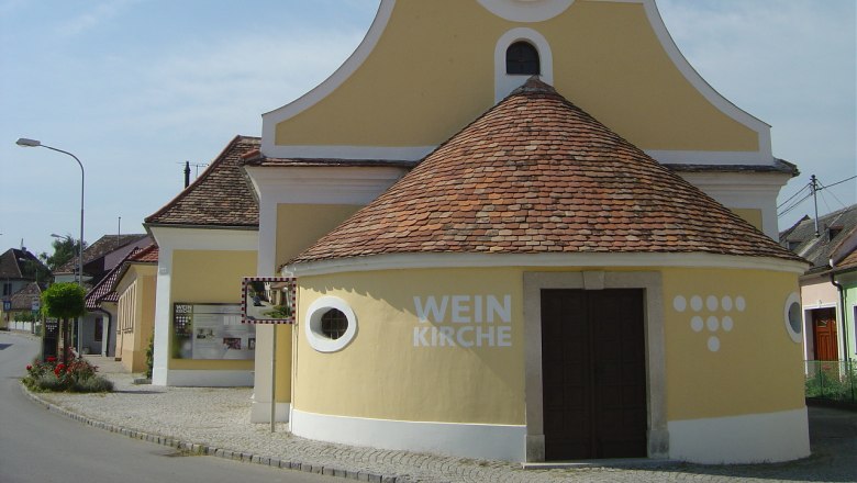 Gelbe Kirche mit Uhrturm und der Aufschrift 'Weinkirche' in einem Dorf.