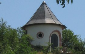Ein runder Wasserturm mit spitzem Dach und Wetterfahne, umgeben von Bäumen und Wiese.