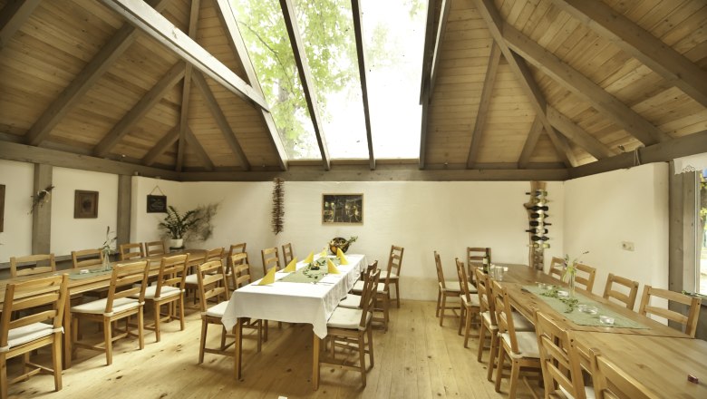 Innenansicht eines rustikalen Raumes mit Holztischen und -stühlen, einem großen Dachfenster und dekorativen Pflanzen.