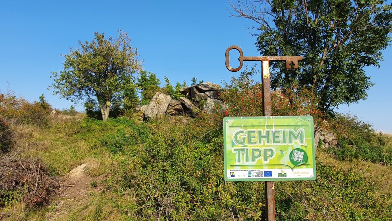 Ein Schild mit der Aufschrift 'Geheimtipp' vor einer natürlichen Landschaft mit Bäumen und Felsen.