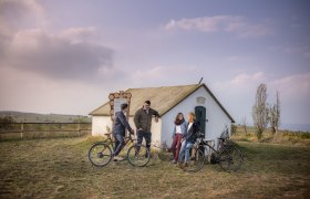 Vier Personen mit Fahrr&auml;dern vor einer kleinen H&uuml;tte in einer l&auml;ndlichen Landschaft.