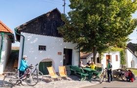 Menschen sitzen auf Bänken vor einem weißen Gebäude mit Fahrrädern in Alberndorf.