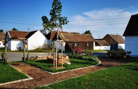 Ein l&auml;ndlicher Hof mit wei&szlig;en Geb&auml;uden, einem kleinen Garten und einem Baum unter blauem Himmel.