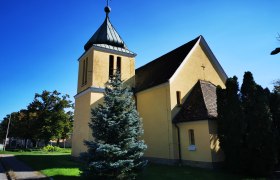 Gelbe Kapelle mit Turm und Kreuz, umgeben von Bäumen, unter blauem Himmel.