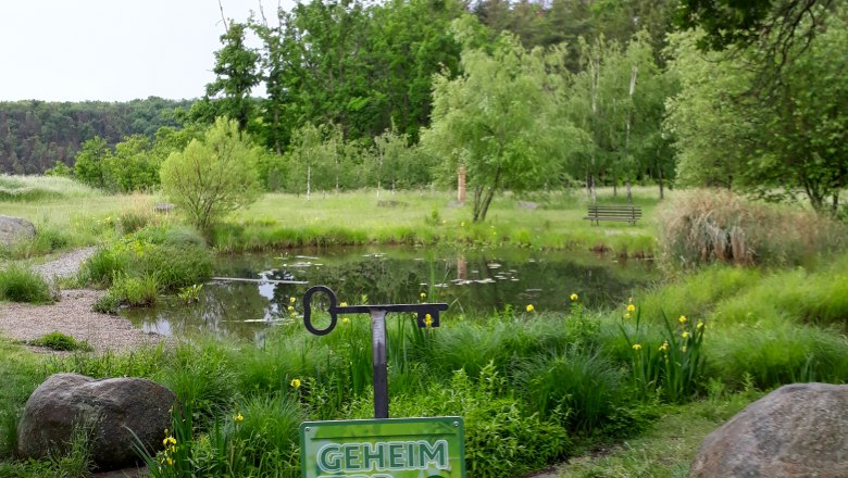Ein grüner Teich mit Bäumen im Hintergrund und einem Schild mit der Aufschrift 'Geheim Tipp' im Vordergrund.