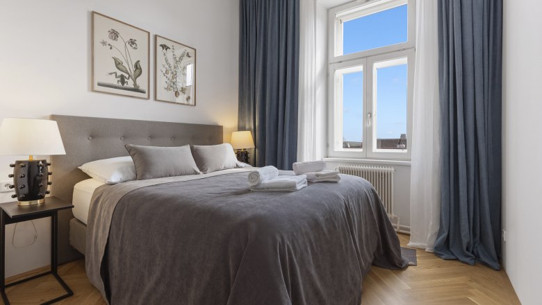 Ein elegantes Schlafzimmer mit einem großen Bett, grauer Bettwäsche, zwei Nachttischen mit Lampen und einem Fenster mit blauem Vorhang.