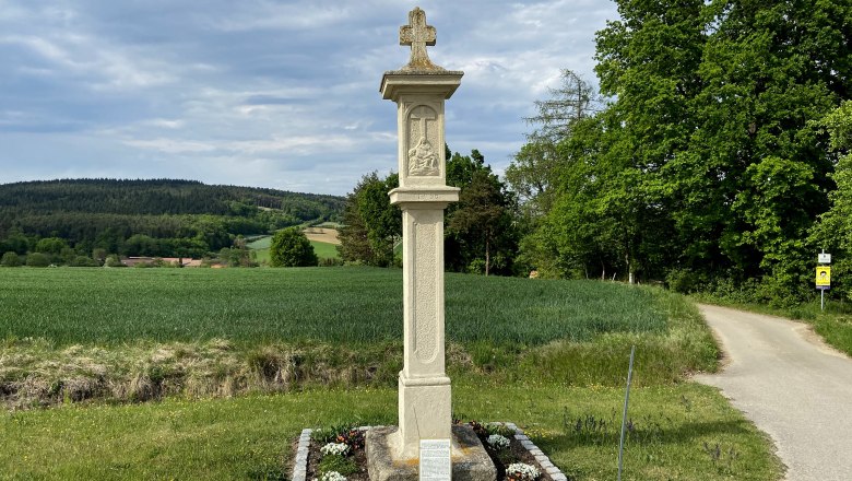 Steinkreuz auf einem Feldweg mit grüner Landschaft im Hintergrund.
