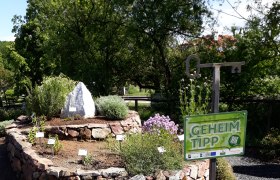 Kräuterspirale aus Steinen mit Pflanzen und einem Schild mit der Aufschrift 'Geheimtipp'.