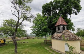 Kleine Kapelle mit rotem Ziegeldach in einer grünen Landschaft, umgeben von Bäumen und Bänken.
