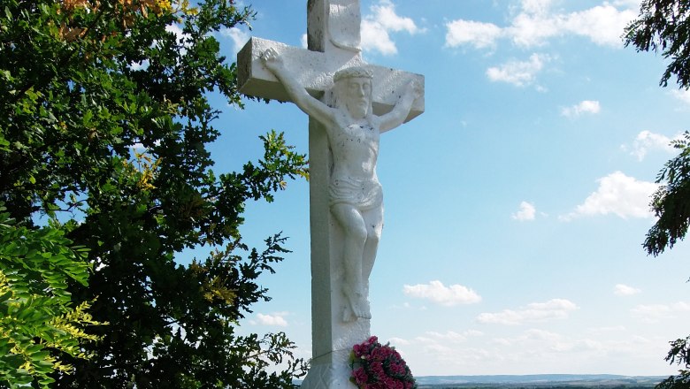 Weißes Steinkreuz mit Jesusfigur und Blumenkranz, umgeben von Bäumen und Landschaft im Hintergrund.