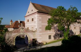 Historisches Gebäude mit rosa Fassade und Torbogen, umgeben von Bäumen.