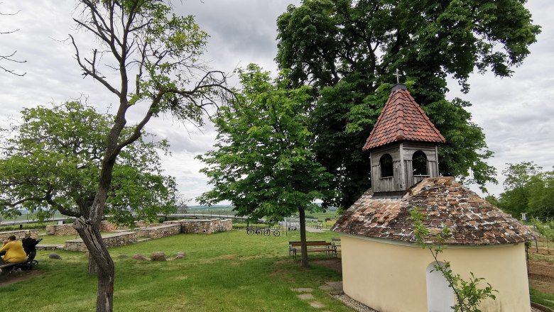 Kleine Kapelle mit rotem Ziegeldach in einer grünen Landschaft, umgeben von Bäumen und Bänken.