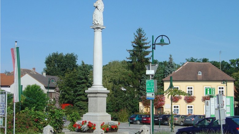 Statue auf einer Säule in einem Platz in Haugsdorf, umgeben von Gebäuden und Bäumen.
