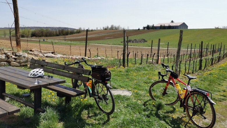 Zwei Fahrräder stehen neben einem Holztisch auf einer Wiese mit Blick auf Felder und ein Gebäude im Hintergrund.