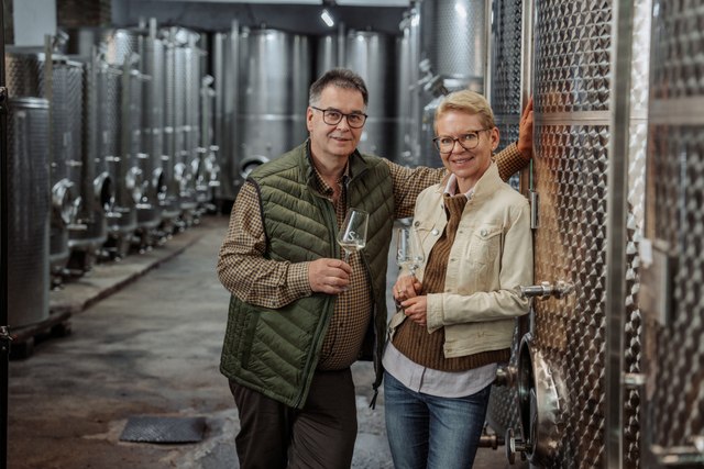 Ein Mann und eine Frau stehen in einer Weinkellerei vor gro&szlig;en Edelstahltanks, beide halten ein Weinglas.