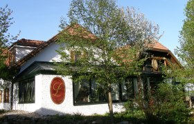 Außenansicht eines traditionellen Gebäudes mit roten Dachziegeln und einem großen L-Logo an der Wand, umgeben von Bäumen und Pflanzen.