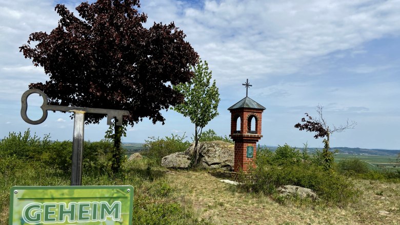 Ein Schild mit der Aufschrift 'Geheim Tipp' vor einem kleinen Backsteinturm mit Kreuz auf einem Hügel.