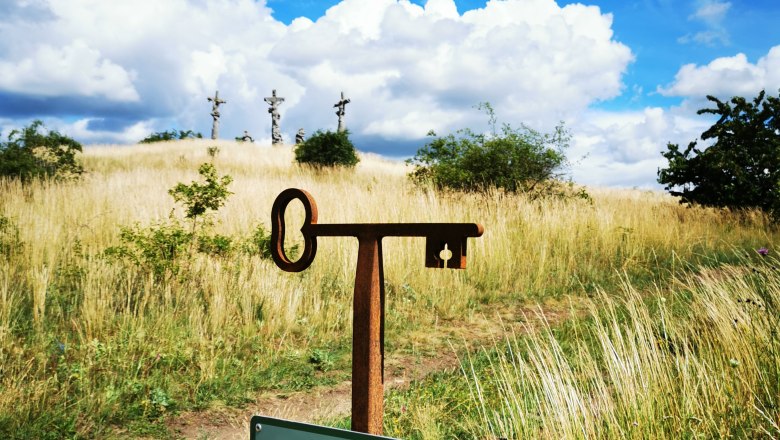Ein Fotopunkt-Schild vor einer Wiese mit drei Kreuzen im Hintergrund unter blauem Himmel mit Wolken.