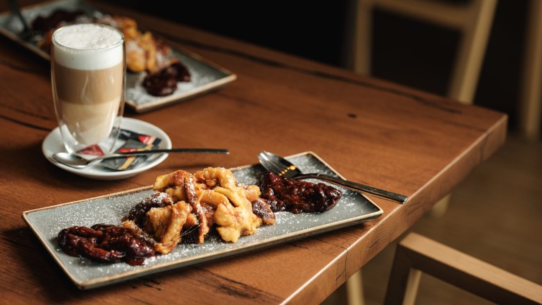 Ein Teller mit Kaiserschmarrn und Pflaumenkompott auf einem Holztisch, daneben ein Glas Latte Macchiato.
