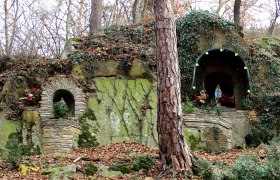 Eine steinerne Grotte mit einer Marienstatue und Pflanzen, umgeben von Bäumen und Laub.