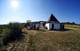 Eine Gruppe von Menschen versammelt sich bei einer kleinen, weißen Hütte mit spitzem Dach auf einer Wiese unter klarem Himmel.