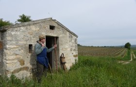 Ein Mann mit Strohhut steht vor einer kleinen Steinhütte in einem Weinberg.