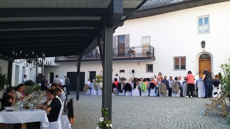 Menschen sitzen an langen Tischen in einem gepflasterten Innenhof bei einer Veranstaltung.