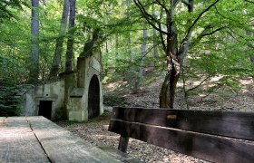 Waldszene mit Brunnstube im Hintergrund und Holzbänken.