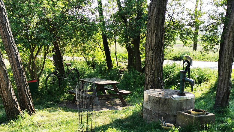 Ein idyllischer Rastplatz mit Tisch, Bank, Fahrrad und Wasserpumpe im Grünen.