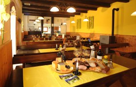 Innenansicht eines gemütlichen Heurigen mit Holztischen, gelben Tischdecken und einer Auswahl an Käse, Brot und Wein auf einem Tisch.