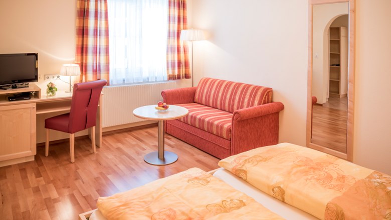 Gemütliches Gästezimmer mit Doppelbett, rotem Sofa, Schreibtisch, Fernseher und großem Spiegel.