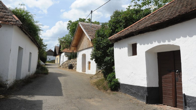 Eine malerische Gasse mit traditionellen Weinkellern in Pillersdorf, umgeben von Bäumen und blauem Himmel.