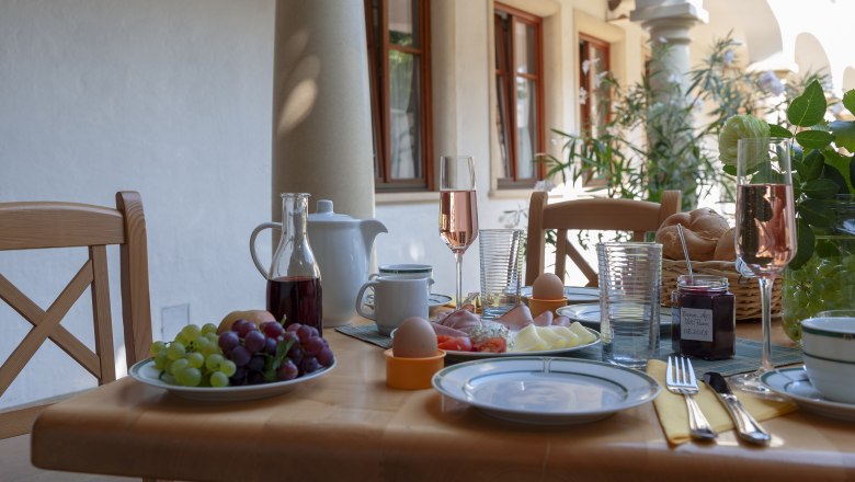 Frühstückstisch im Freien mit Weintrauben, Schinken, Käse, Eiern, Brötchen und Sektgläsern.