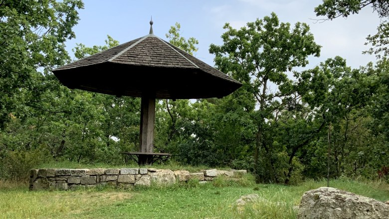 Ein hölzerner Pavillon mit spitzem Dach in einer grünen, bewaldeten Umgebung.