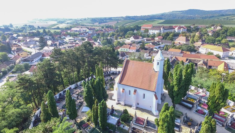 Luftaufnahme einer Kirche mit rotem Dach, umgeben von einem Friedhof und Bäumen, in einer ländlichen Umgebung mit Häusern und Hügeln im Hintergrund.