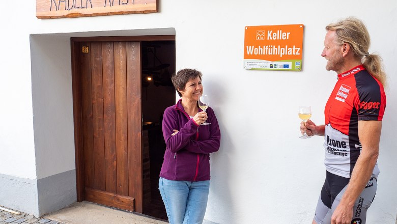 Zwei Personen stehen vor einem Gebäude mit der Aufschrift 'Radler-Rast' und 'Keller Wohlfühlplatz', sie halten Weingläser in der Hand.