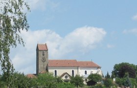 Kirche mit Turm und rotem Dach in Hohenwarth, umgeben von Bäumen und blauem Himmel.