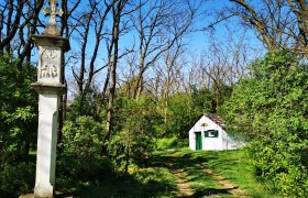 Ein steinernes Kreuz und ein kleines weißes Gebäude in einem Waldgebiet bei sonnigem Wetter.