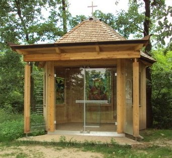Kleine Holzkapelle mit Glasfront und Kreuz auf dem Dach, umgeben von B&auml;umen.