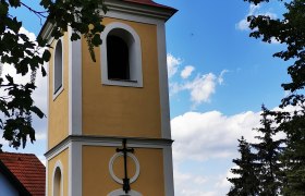 Glockenturm in Ragelsdorf vor blauem Himmel und Bäumen.