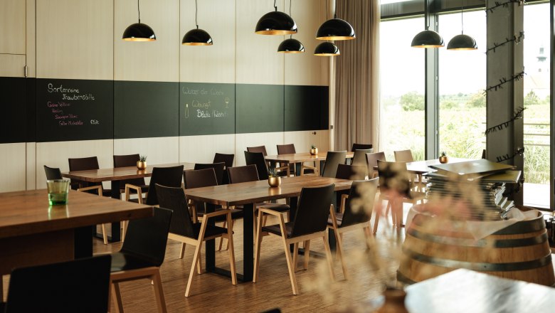 Modernes Restaurant mit Holztischen, schwarzen St&uuml;hlen und gro&szlig;en Fenstern. Schwarze H&auml;ngelampen und eine Tafel an der Wand.