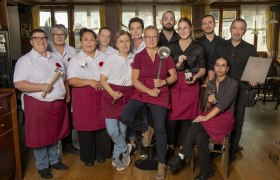 Ein Team von Restaurantmitarbeiterinnen und Mitarbeitern posiert in Arbeitskleidung mit K&uuml;chenutensilien in einem Speiseraum.